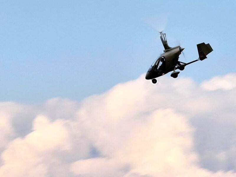 Gyrocoptère noir volant devant un ciel bleu avec des nuages blancs en arrière-plan.