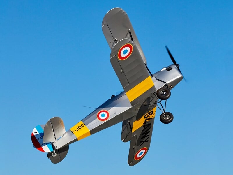 Avion vintage gris avec cocardes françaises en vol incliné sur fond de ciel bleu.