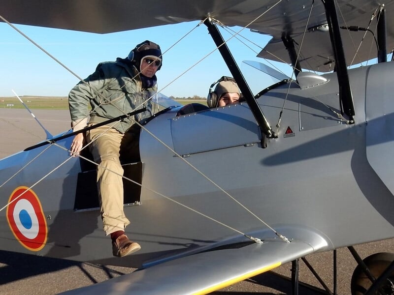 Personne montant dans un avion biplan gris avec cocardes françaises sur piste d'aérodrome.