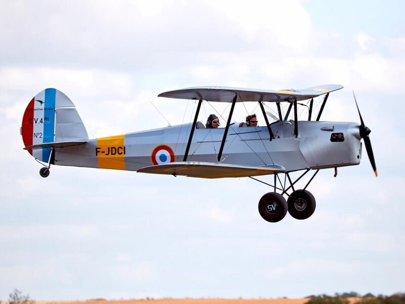 Avion biplan gris avec cocardes françaises volant au-dessus d'un champ sous un ciel clair.