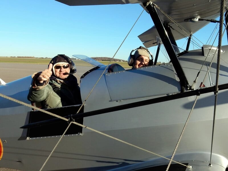 Deux personnes souriantes dans un avion biplan gris avec cocardes françaises sur piste.