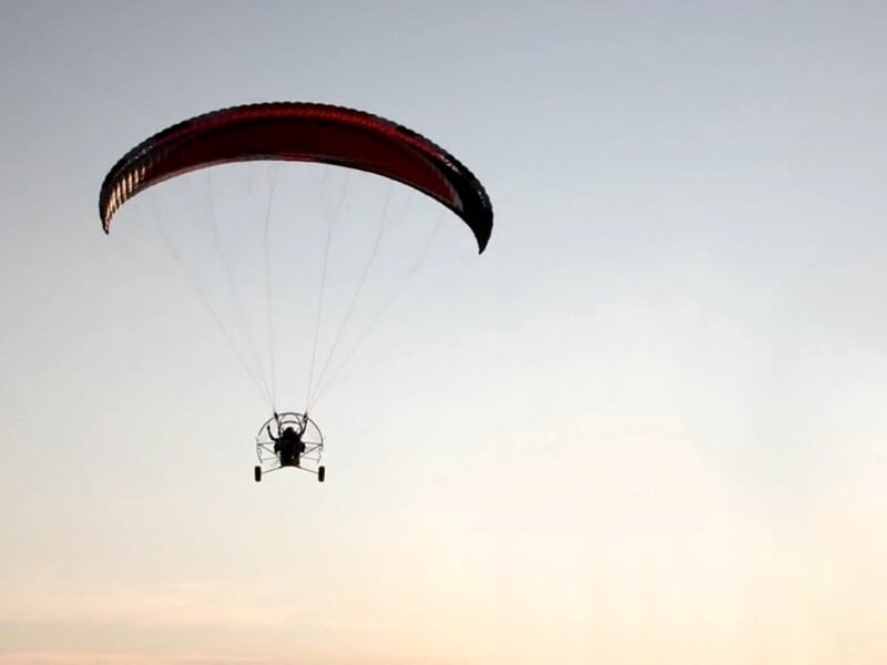 Paramoteur volant au coucher du soleil avec voile rouge et ciel clair en arrière-plan.