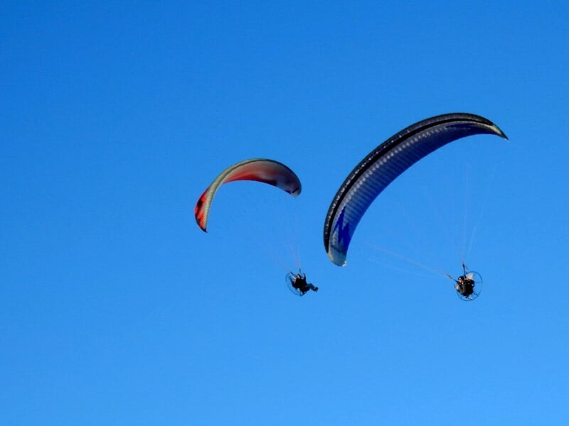 Deux paramoteurs en vol avec voiles colorées rouge et bleue sur fond de ciel bleu.