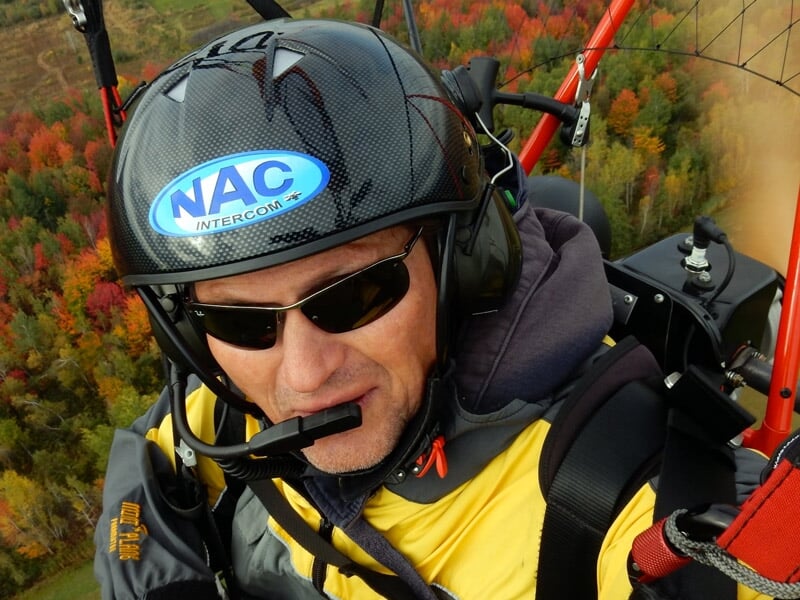 Homme en casque et lunettes de soleil en vol en paramoteur au-dessus d'une forêt colorée.