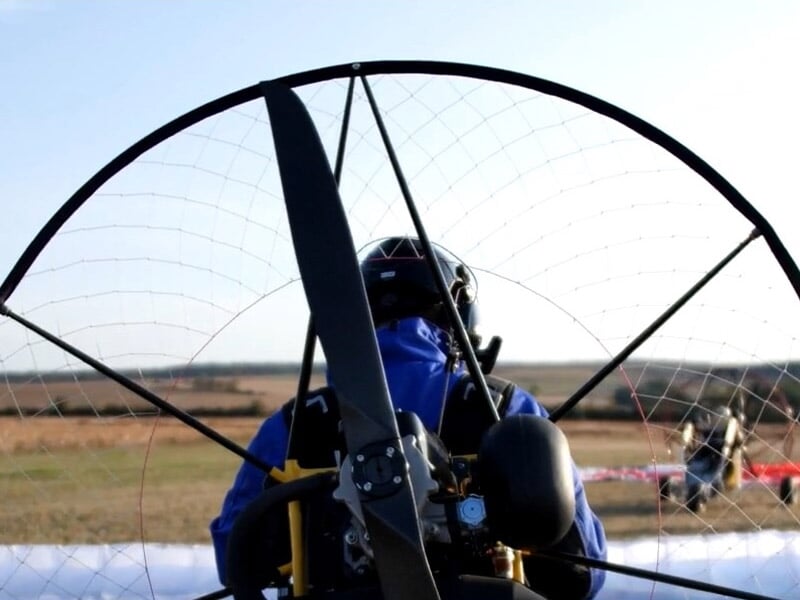 Vue arrière d'un pilote en paramoteur avec moteur et hélice visibles au sol.