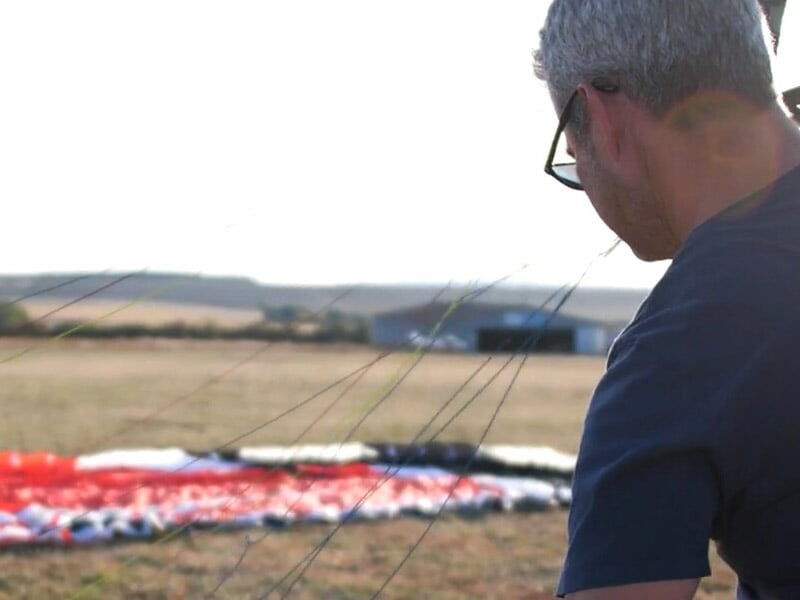 Homme préparant un parapente rouge et blanc sur un terrain d'aérodrome.