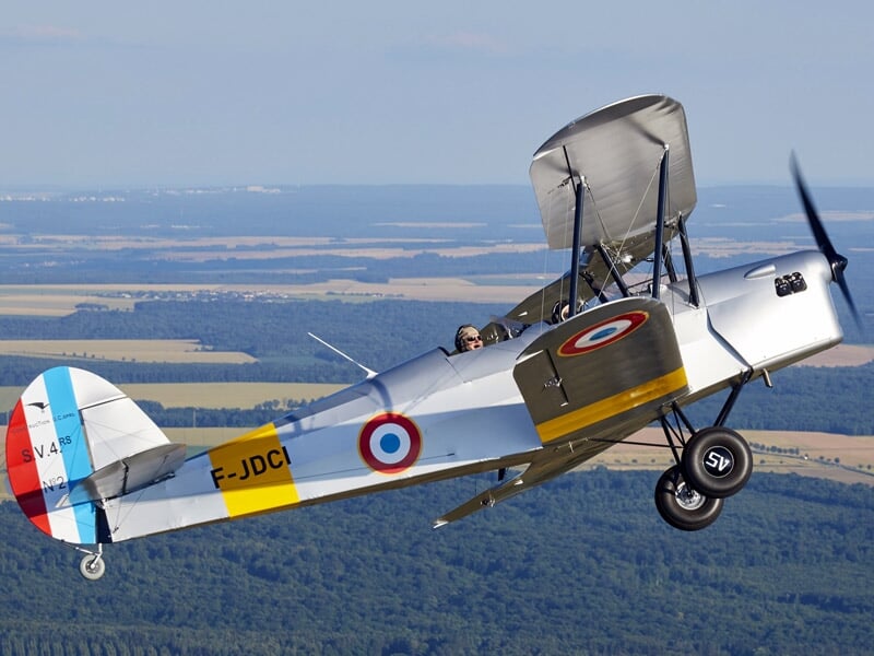 Avion biplan gris avec cocardes françaises volant au-dessus d'un paysage rural.