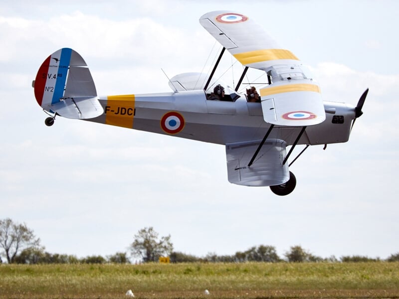 Avion biplan gris avec cocardes françaises volant au-dessus d'un champ sous ciel clair.
