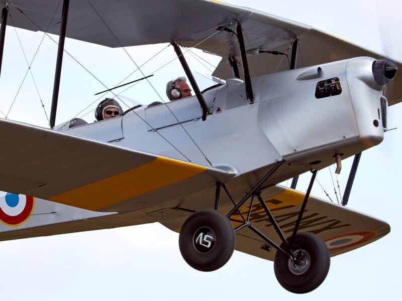 Deux personnes dans un avion biplan gris avec cocardes françaises en vol.