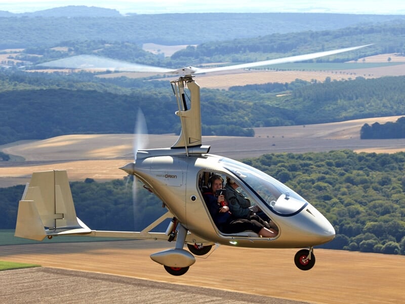 Gyrocoptère gris volant au-dessus d'un paysage de campagne avec ciel clair.