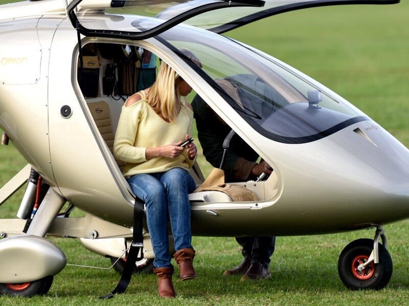 Femme assise dans un gyrocoptère gris au sol, prête à décoller sur une piste verte.