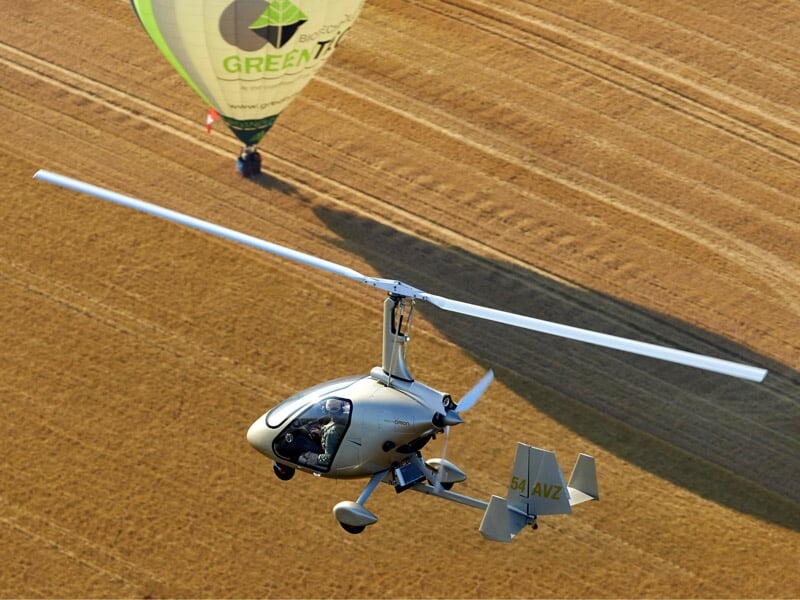 Gyrocoptère gris volant au-dessus d'un champ avec une montgolfière verte en arrière-plan.