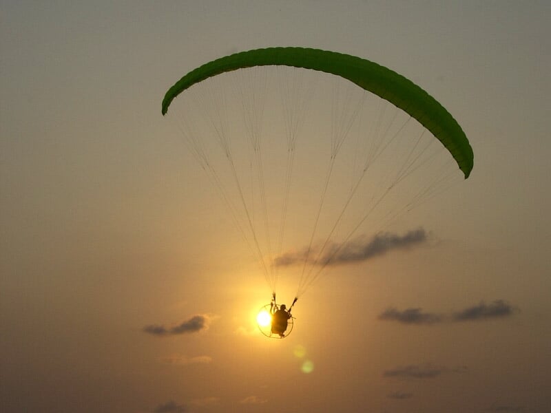 Paramoteur en vol au coucher du soleil avec voile verte et ciel orangé.