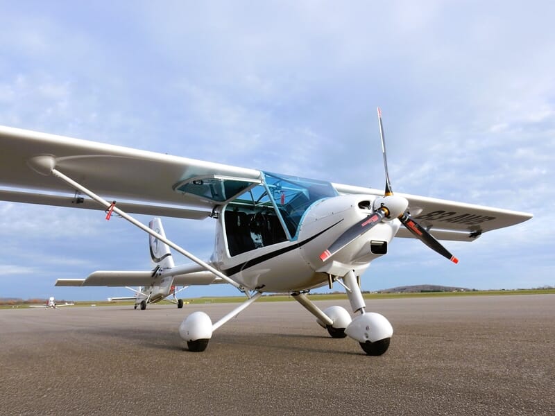 Avion ULM blanc sur piste avec hélice, ailes déployées et ciel nuageux en arrière-plan.
