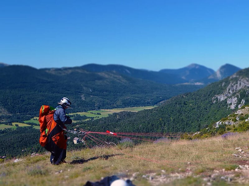 Personne préparant son équipement de parapente sur une colline avec vue sur les montagnes et la vallée en arrière-plan.