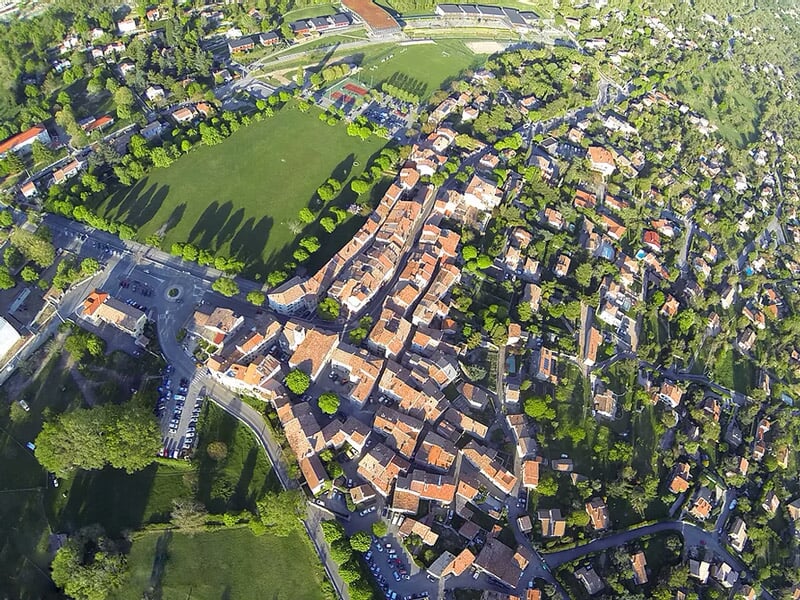 Vue aérienne d'un village avec des maisons, des rues et un terrain de sport entouré de verdure.