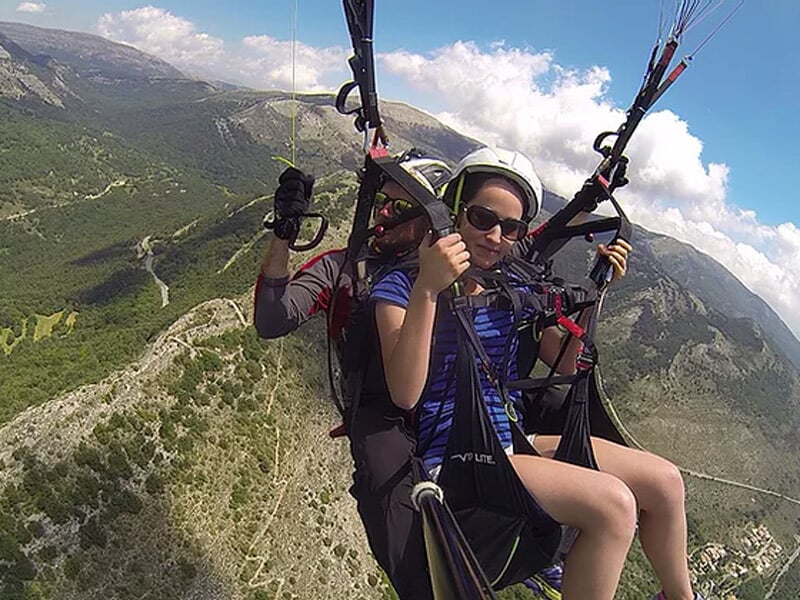 Deux personnes en vol en parapente, l'une tenant les commandes, survolant une région montagneuse verdoyante.