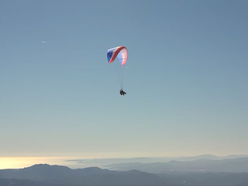 Parapente rouge et blanc en vol au-dessus des montagnes avec un ciel bleu clair et un horizon lointain.