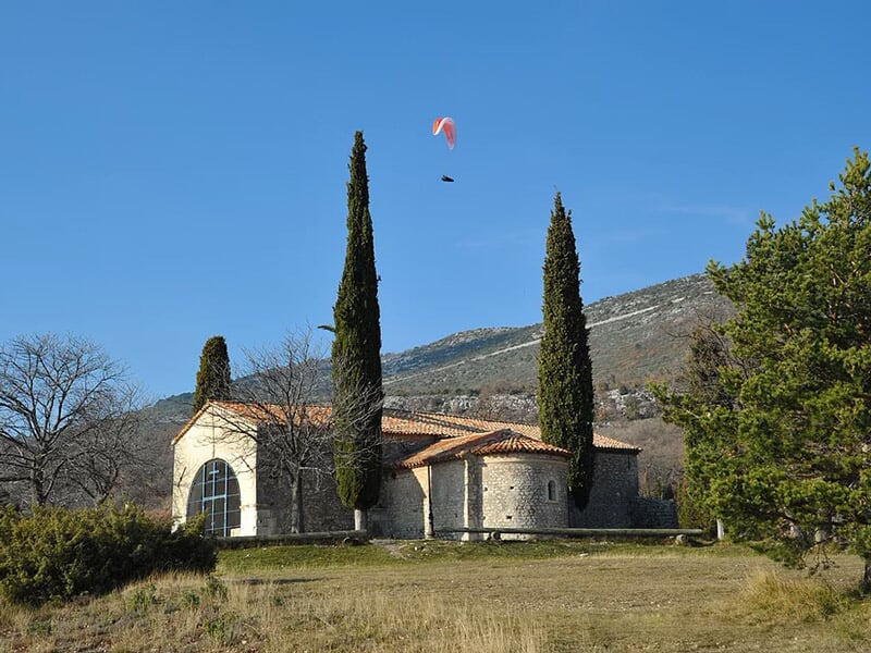 Petit bâtiment en pierre entouré d'arbres et de collines, avec un parapente rouge visible dans le ciel.