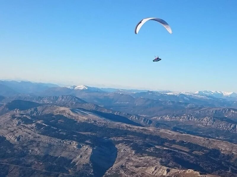 Parapente blanc volant au-dessus d'un paysage montagneux avec des sommets enneigés au loin sous un ciel bleu.