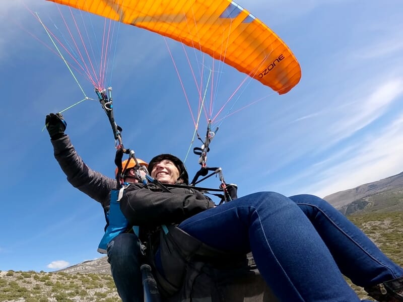 Deux personnes en parapente avec une voile orange survolant un paysage montagneux sous un ciel bleu clair.