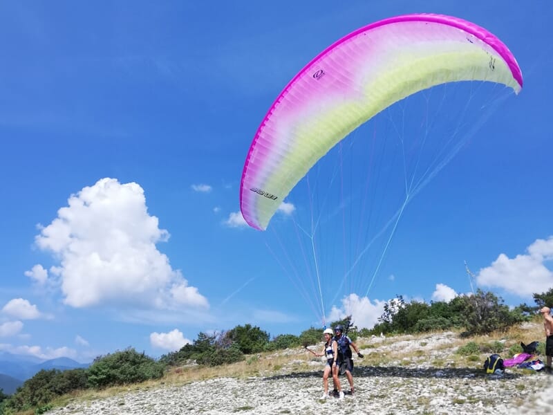 Parapente rose et blanc au sol avec deux personnes prêtes à décoller sur un terrain rocheux et ciel bleu avec nuages.