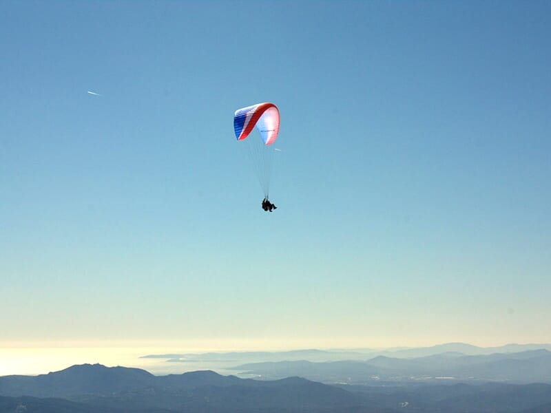 Parapente tricolore rouge, blanc et bleu en vol au-dessus de montagnes sous un ciel dégagé et lumineux.
