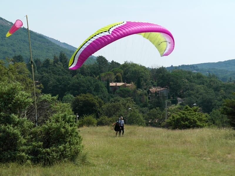 Personne en parapente avec voile rose et jaune survolant une prairie verte avec arbres et collines en arrière-plan.