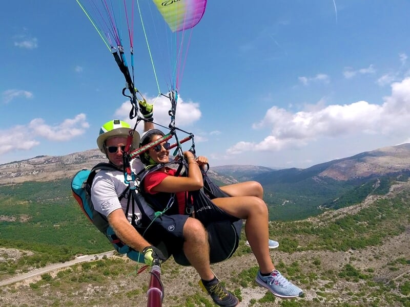 Deux personnes en parapente avec voile rose et jaune volant au-dessus d'un paysage montagneux sous un ciel partiellement nuageux.