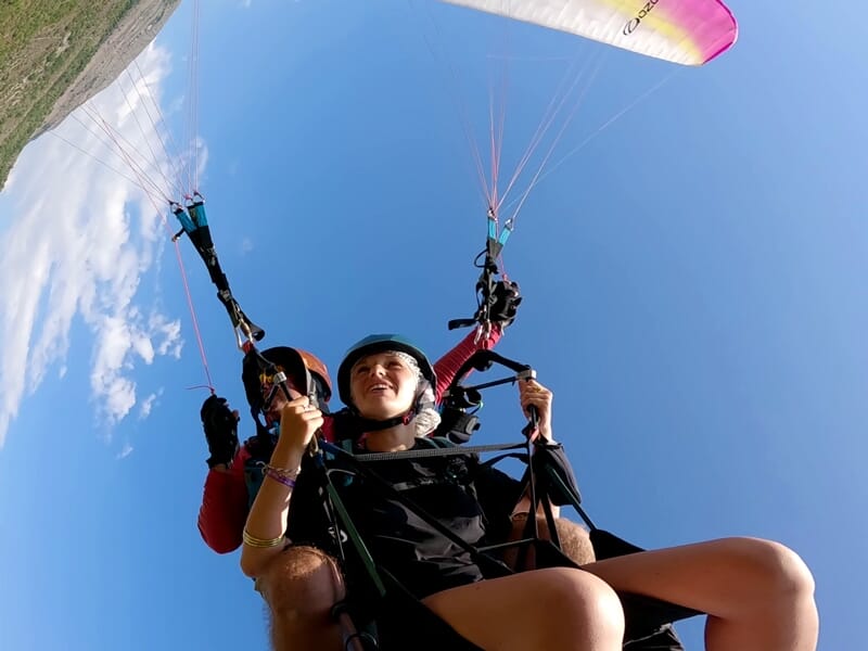 Jeune femme souriante en parapente avec casque noir et voile rose et jaune, ciel bleu avec quelques nuages.