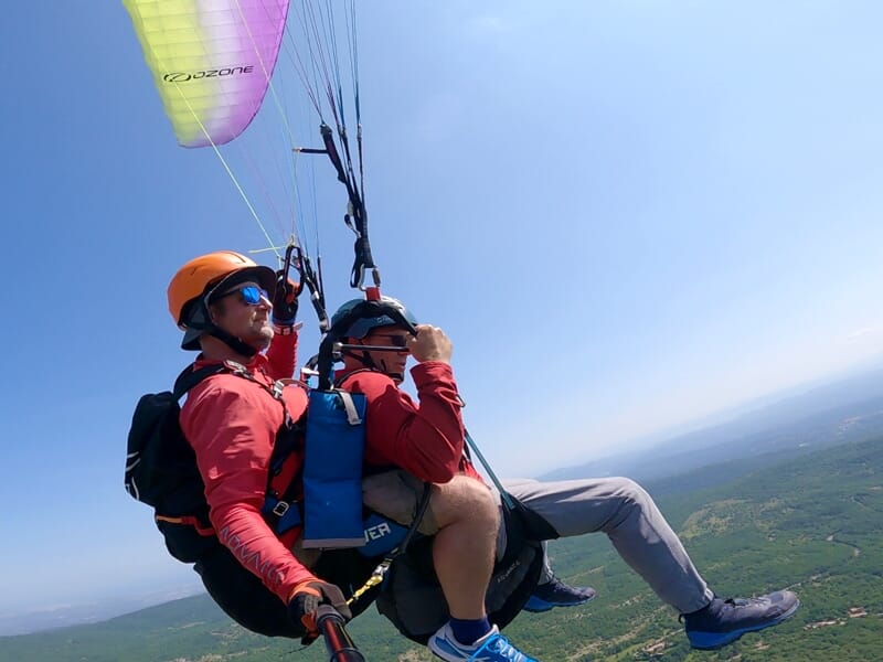 Homme en parapente avec casque orange et lunettes, accompagné d'une passagère, survolant un paysage vert sous un ciel clair.