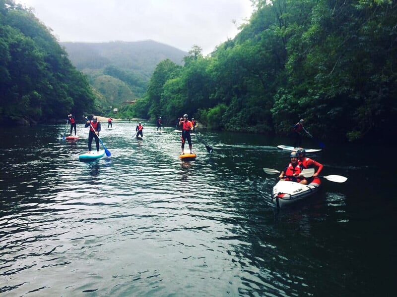 Groupe de personnes en combinaison rouge pratiquant le paddle sur une rivière entourée d'arbres verts et collines brumeuses.