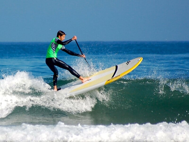 Surfeur en combinaison noire et verte effectuant une figure sur une planche de paddle dans une vague bleue de l'océan.