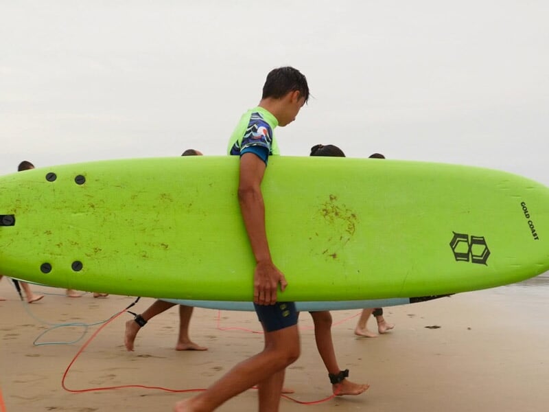Jeune garçon portant une combinaison bleue et verte portant une grande planche de surf verte sur une plage de sable.