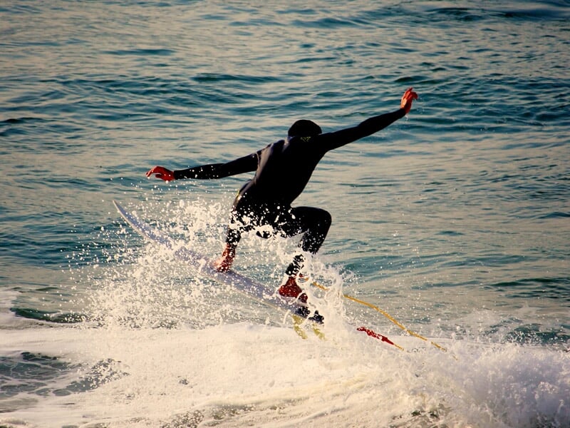 Surfeur en combinaison noire sautant sur une planche de surf sur une vague, éclaboussant l'eau de mer bleue.