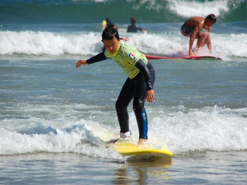 Enfant en combinaison noire et jaune debout sur une planche de surf jaune dans une petite vague sur une plage.