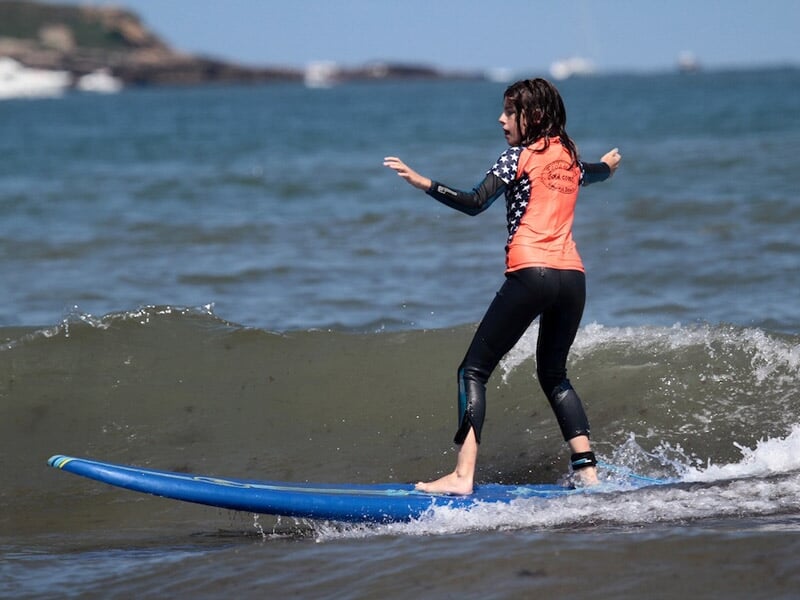 Jeune surfeuse en combinaison noire et orange debout sur une planche bleue surfant une petite vague sur l'océan.
