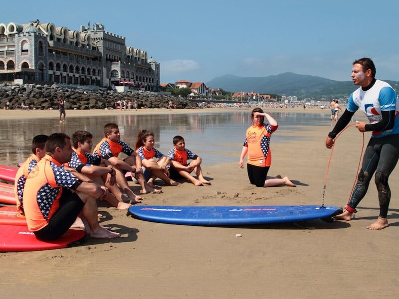 Groupe d'enfants en combinaison orange assis sur la plage écoutant un instructeur debout près de planches de surf bleues.
