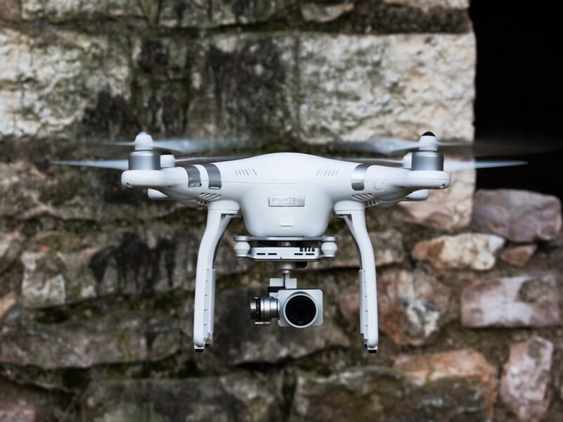 Drone blanc en vol stationnaire devant un mur en pierre, équipé d'une caméra sous le corps principal.