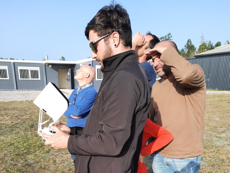 Groupe de personnes en extérieur, un homme tient une télécommande pour piloter un drone dans le ciel.