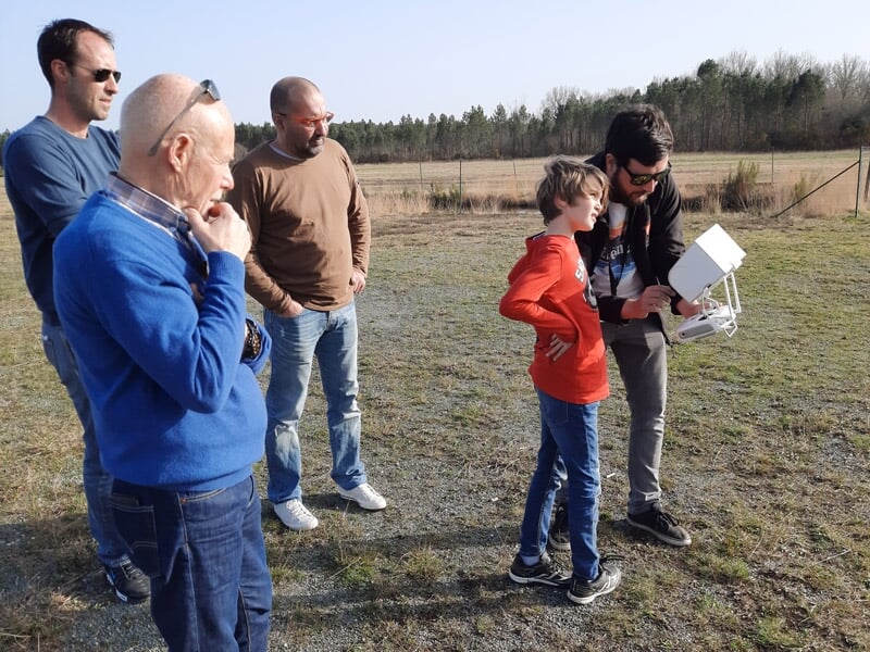Quatre personnes debout sur un terrain herbeux, un homme montre à un enfant comment piloter un drone.