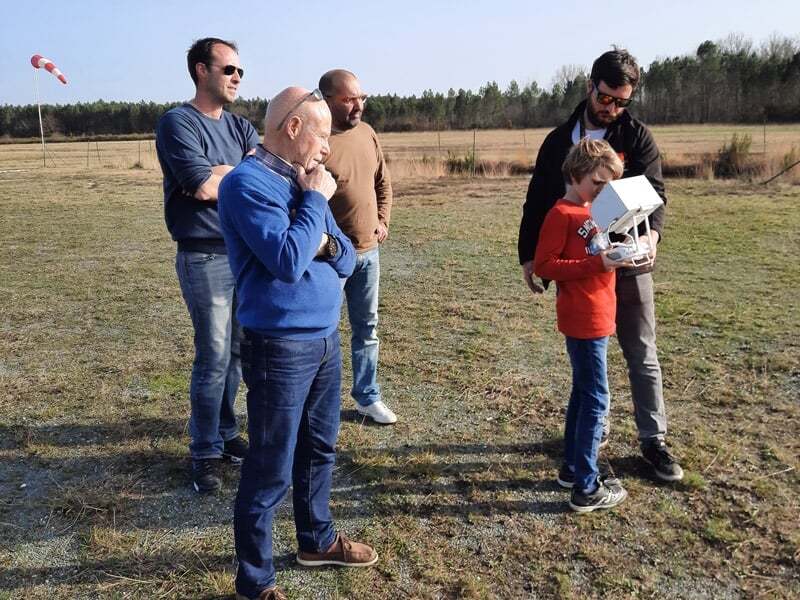 Groupe de quatre personnes sur un terrain, un enfant manipule une télécommande de drone avec aide.