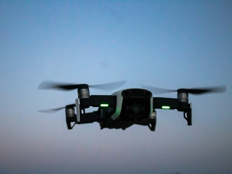 Drone noir en vol au crépuscule, ses hélices en mouvement et lumières vertes allumées visibles.