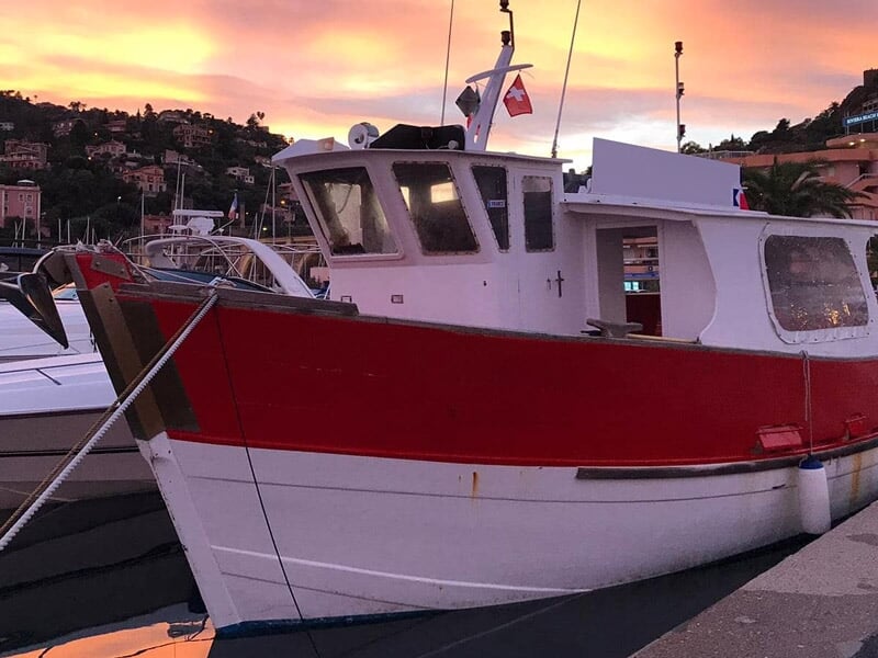 Bateau de pêche rouge et blanc amarré dans un port au coucher du soleil avec des collines en arrière-plan.