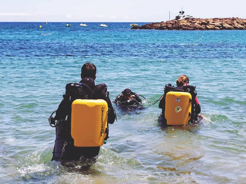 Trois plongeurs avec des bouteilles jaunes marchant dans l'eau peu profonde vers la mer.