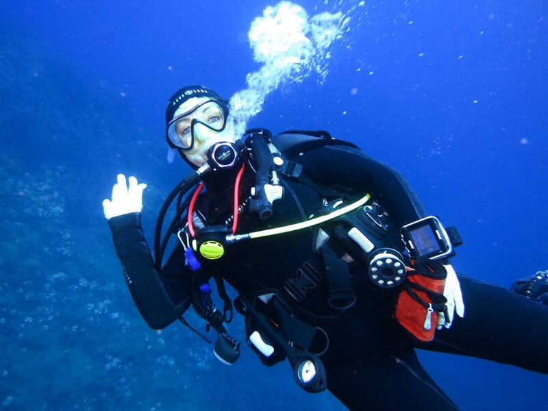 Plongeur souriant en combinaison noire fait un signe de la main sous l'eau bleue claire.