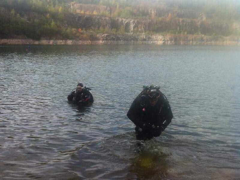 Deux plongeurs en combinaison noire sortent de l'eau dans un lac entouré de falaises et de végétation.