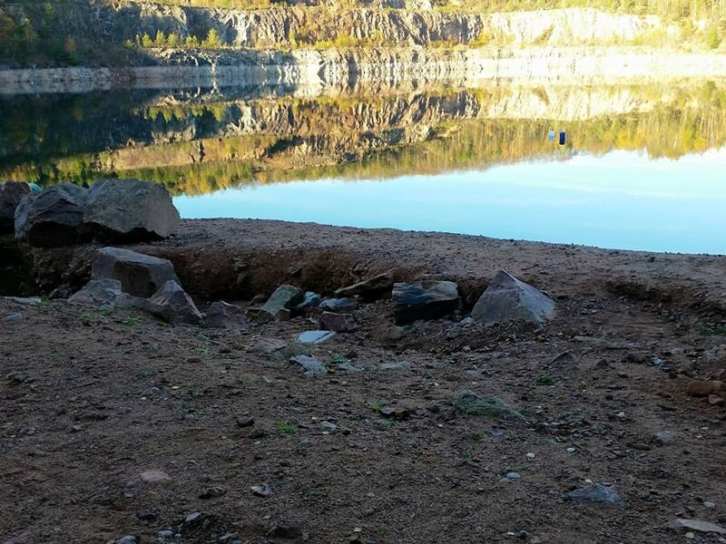 Rivage rocheux et terreux au bord d'un lac calme reflétant les falaises et les arbres environnants.