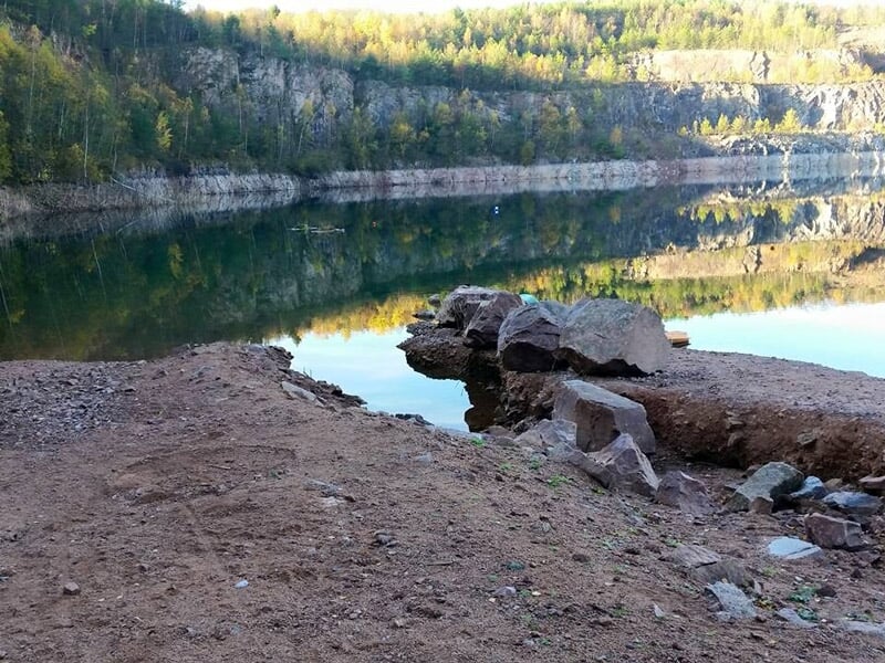 Vue du rivage avec des rochers et un lac paisible entouré de falaises et de forêts en arrière-plan.