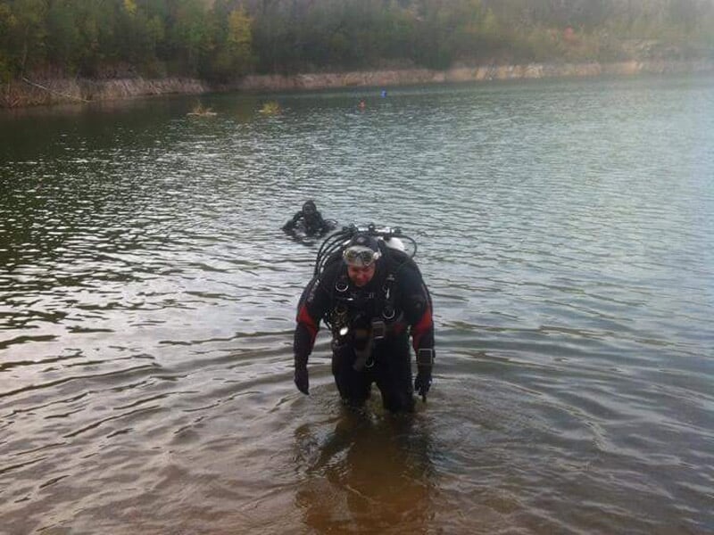 Plongeur en combinaison noire sortant de l'eau dans un lac entouré de falaises et de végétation.